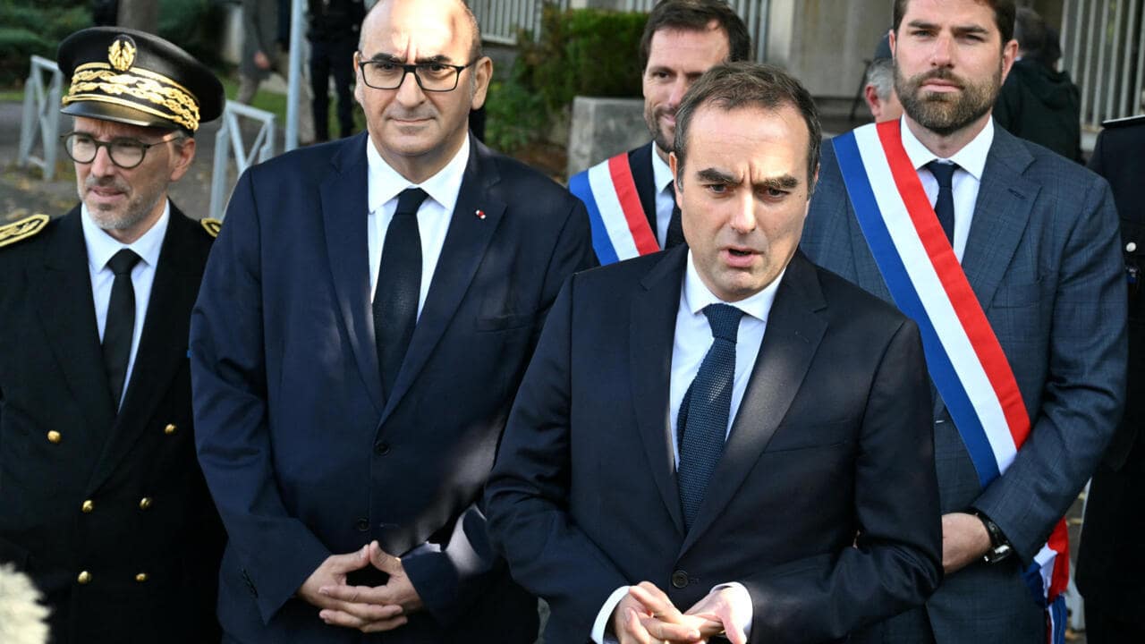 Plusieurs hommes en costume, dont deux au premier plan, devant un bâtiment, lors d'un événement lié à un changement de gouvernement